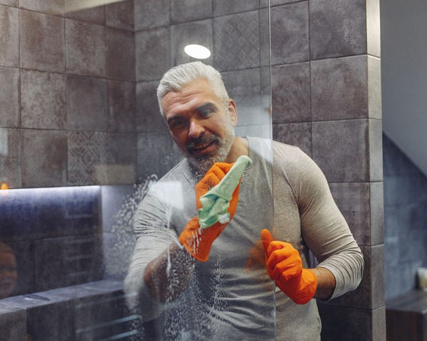 Grey haired male with beard in rubber gloves cheerfully cleaning shower glass shield