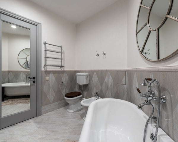 White bathtub with faucet at tiled wall placed near toilet and bidet in contemporary bathroom with mirror with reflection on door